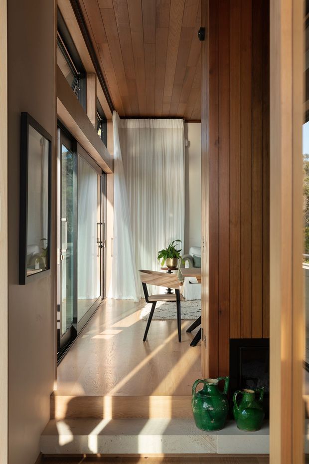 A warm, narrow interior passage or entry space leading into a compact dining and living area. The space features extensive floor-to-ceiling glazing with sheer curtains, a timber-lined ceiling, vertical timber wall panelling, pale timber flooring, and a small mid-century-style dining table and chairs. Strong sunlight creates long shadows across the floor and steps, while green ceramic vessels sit in the foreground.