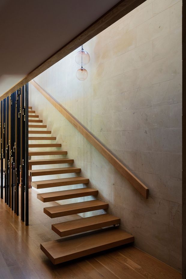 Interior view of a contemporary stairway with floating solid-wood treads, a matching timber handrail, and a pale stone or concrete-block feature wall. A dark vertical slatted screen borders the stair, while two spherical pendant lights hang beneath the angled ceiling. Warm timber flooring and subdued natural tones create a refined, dramatic atmosphere.