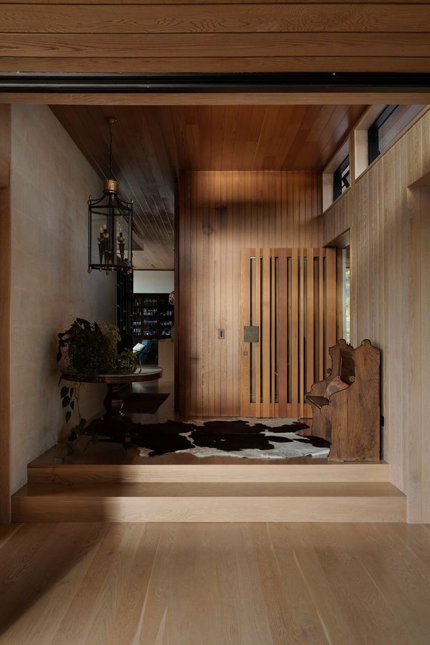 A warm, wood-lined residential entry hall viewed from a lower level, with broad timber flooring, a raised landing, vertical timber wall panelling, a slatted timber front door, and clerestory glazing. A sculptural carved timber bench, dark cowhide rug, console table with plants, and hanging lantern add character. The hallway leads toward a darker living area beyond.