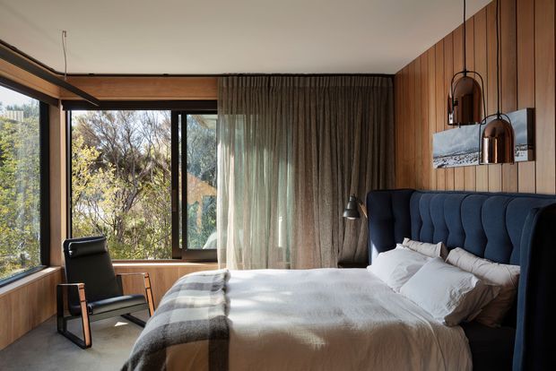 A warm, sophisticated bedroom with a large upholstered bed, layered white bedding, and a dark navy scalloped headboard. Full-height timber wall panelling wraps the room, while expansive corner glazing overlooks trees. Sheer curtains soften the natural light, and a black leather lounge chair, bedside lighting, and suspended pendant lights complete the refined interior.