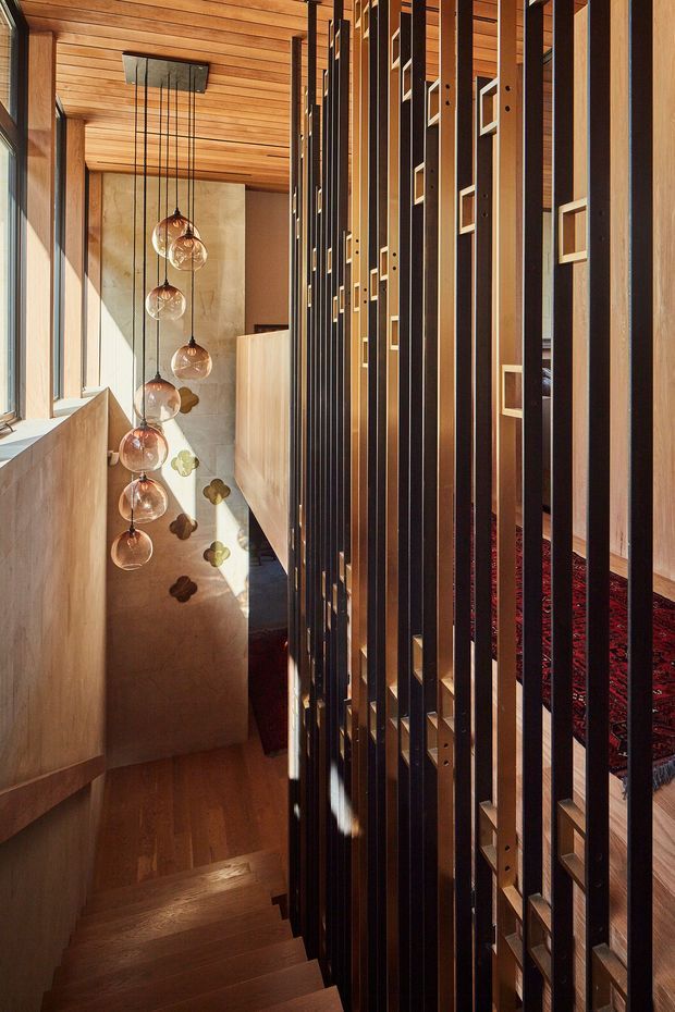 A dramatic interior stairwell viewed from above, with a timber staircase descending beside a tall decorative screen of closely spaced dark metal and timber vertical slats. The stair hall features warm timber wall and ceiling lining, pale plaster or concrete walls, large glazing, and a cascading cluster of spherical pendant lights. A patterned dark red rug is visible through the screen on the adjacent upper-level area. Strong sunlight creates pronounced shadows across the walls and stairs.