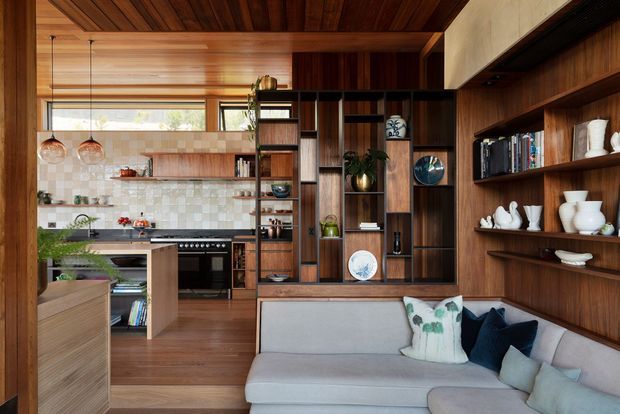 Warm open-plan living and kitchen interior with extensive dark timber joinery, a built-in shelving and display wall, timber ceiling, pale sectional sofa, and a compact kitchen with island, range and patterned tiled splashback. Pendant lights and carefully styled shelving create a refined mid-century-inspired atmosphere.
