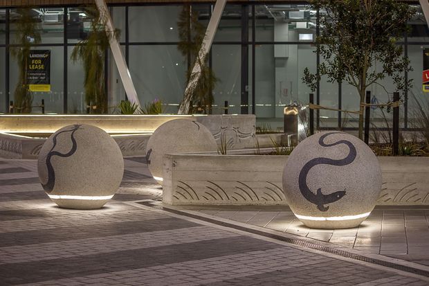 Night-time view of a contemporary commercial courtyard or plaza featuring three illuminated spherical concrete sculptures with dark abstract markings, integrated concrete planter benches, patterned paving, linear lighting, trees, and a glazed modern building facade in the background. A leasing sign is visible through the glazing.