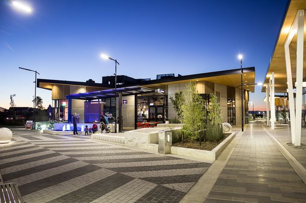 A contemporary commercial building and landscaped forecourt at dusk, featuring a low flat roof, broad glazing, illuminated interiors, a covered pedestrian arcade with white columns, and patterned geometric paving. Small planted beds and concrete seating define the public entry area.