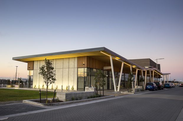 Exterior architectural photograph of a contemporary low-rise commercial building at dusk. The building features broad flat roof planes with deep overhangs, warm recessed soffit lighting, large glazed walls, pale concrete or panel-clad walls, timber-toned soffit areas, and distinctive angled white structural columns. A paved forecourt, landscaping, and parked cars are visible.