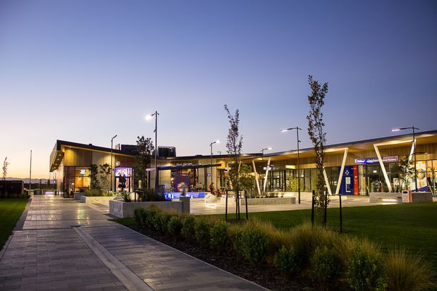 Dusk exterior view of a contemporary single-storey commercial complex with broad glazed facades, projecting flat rooflines, warm illuminated interiors, landscaped lawns, ornamental grasses, young trees, and a paved pedestrian walkway leading through the site.
