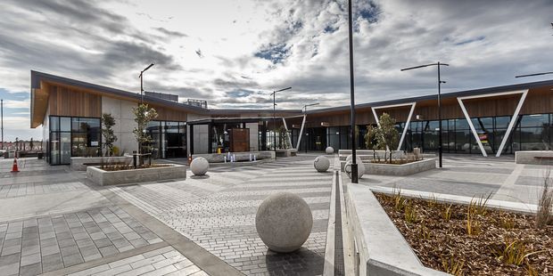 Wide exterior view of a contemporary low-rise commercial or civic complex arranged around a paved public plaza. The buildings feature extensive floor-to-ceiling glazing, timber-lined soffits and facade sections, pale masonry or concrete walls, and distinctive angled white structural frames. The plaza includes patterned paving, raised planter beds, sculptural concrete spheres, slim modern light poles, and outdoor seating elements under a dramatic overcast sky.