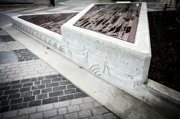 A low concrete architectural structure or raised planter-like feature with decorative relief detailing, topped with dark timber or metal inset panels. It sits beside a paved surface composed of contrasting light and dark rectangular pavers. The image focuses on the built outdoor element and its textured, ornamental finish.