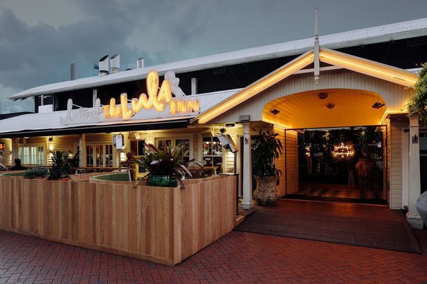 Exterior dusk view of a low-rise hospitality venue with a prominent illuminated sign, covered entrance, timber-clad outdoor dining enclosure, brick paving, and warm interior lighting visible through the open frontage. The building has a long pitched roof and a welcoming restaurant or inn-like facade.