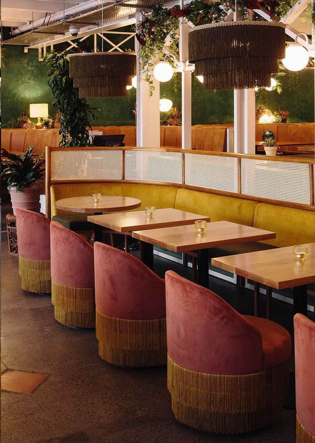 A richly styled restaurant interior with rows of curved upholstered banquettes and dining chairs, warm timber tables, woven cane privacy screens, brass-toned fringe detailing, globe pendant lights, and abundant hanging greenery. Deep green walls, terracotta-pink upholstery, mustard seating, and polished concrete flooring create a dramatic, retro-inspired hospitality setting.