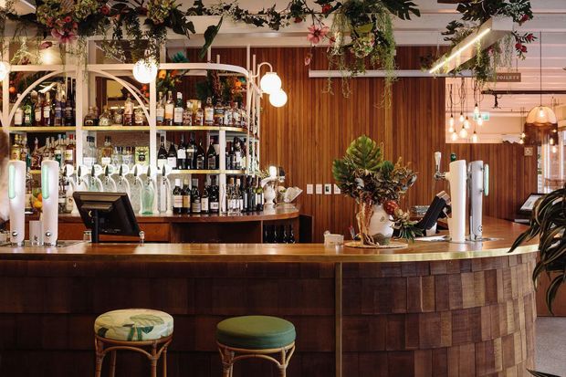 A richly styled commercial bar interior with a curved dark timber bar front, timber wall panelling, extensive bottle shelving, brass-toned and globe lighting, and abundant hanging greenery and floral arrangements. The space has a warm, botanical, hospitality-focused atmosphere with visible bar equipment and seating.