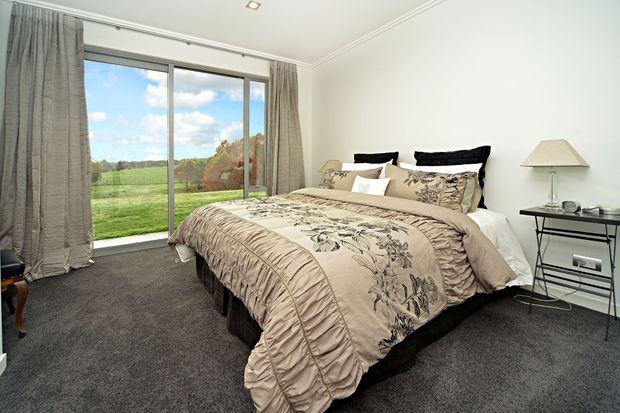 A bright, traditionally styled bedroom with a large bed dressed in beige floral bedding, dark upholstered headboard cushions, grey carpet, full-height grey curtains, and a slim black bedside table. Large sliding glass doors open to a green rural landscape.
