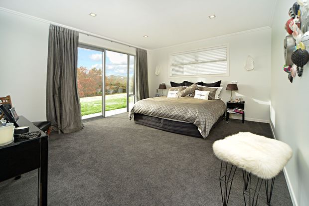 A bright, spacious contemporary bedroom with a large upholstered bed, grey quilted bedding, dark grey carpet, white walls, and floor-to-ceiling sliding glass doors opening onto a lawn and garden. A wide window with white blinds, heavy grey curtains, bedside tables, a compact black desk or console, and a white furry stool complete the room. Decorative sculptural wall pieces add a small accent on the right.