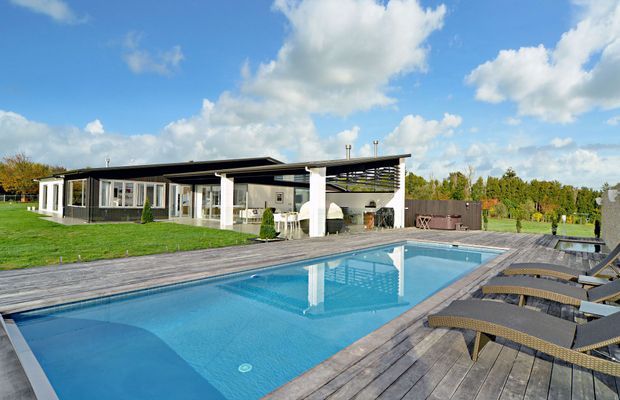 A low, modern single-storey residence with extensive glazing and a broad flat roof, set beside a large rectangular swimming pool. The pool is surrounded by a weathered timber deck with multiple outdoor loungers, while a covered outdoor entertaining area and landscaped lawn extend around the house. The image has a bright, open, resort-like atmosphere under a blue sky.