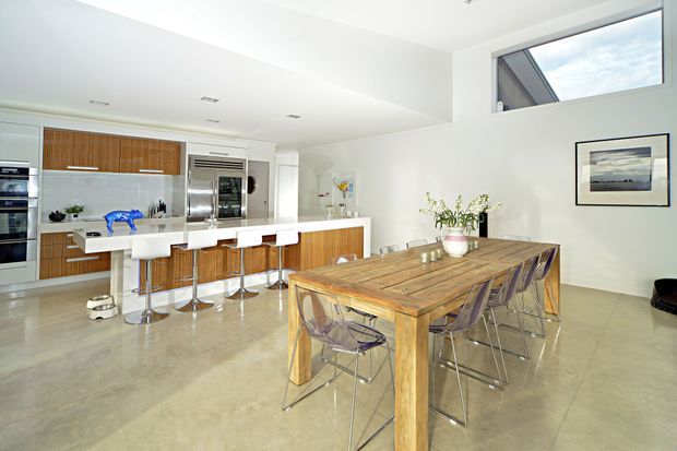 Bright open-plan kitchen and dining area with white walls and ceiling, warm timber cabinetry, a large kitchen island with white stools, stainless-steel appliances, polished concrete flooring, and a substantial timber dining table with transparent modern chairs. High-level glazing brings in natural light, while framed artwork and a vase provide restrained decoration.