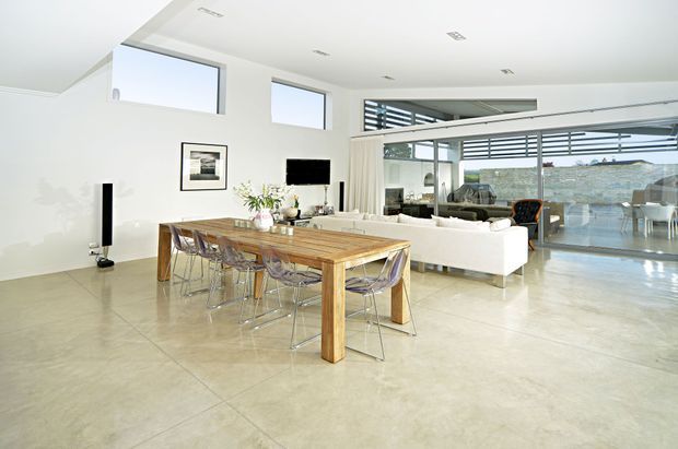 Bright open-plan contemporary living and dining space with polished concrete flooring, white walls, a large timber dining table with clear chairs, and a white sectional sofa. Full-height glazing opens toward an outdoor terrace and distant landscape, while high windows and recessed ceiling lights create an airy atmosphere.