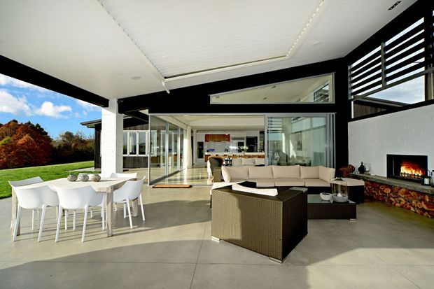 A bright, expansive open-plan living and entertaining area with floor-to-ceiling glazing opening onto a lawn. The space features a white dining table and chairs, woven outdoor-style lounge furniture, a contemporary fireplace with a colourful stone-clad hearth, polished concrete flooring, a white paneled ceiling, and black-framed architectural screens and glazing. The kitchen and additional living space are visible beyond the main seating area.