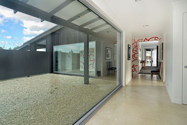 A bright contemporary interior hallway beside a large floor-to-ceiling glazed opening overlooking a gravel courtyard. The space features smooth light flooring, white walls and ceiling, dark metal-framed glazing, and a covered outdoor structure with dark vertical cladding. Red graphic artwork is visible along the hallway, with a glimpse of a furnished room beyond.