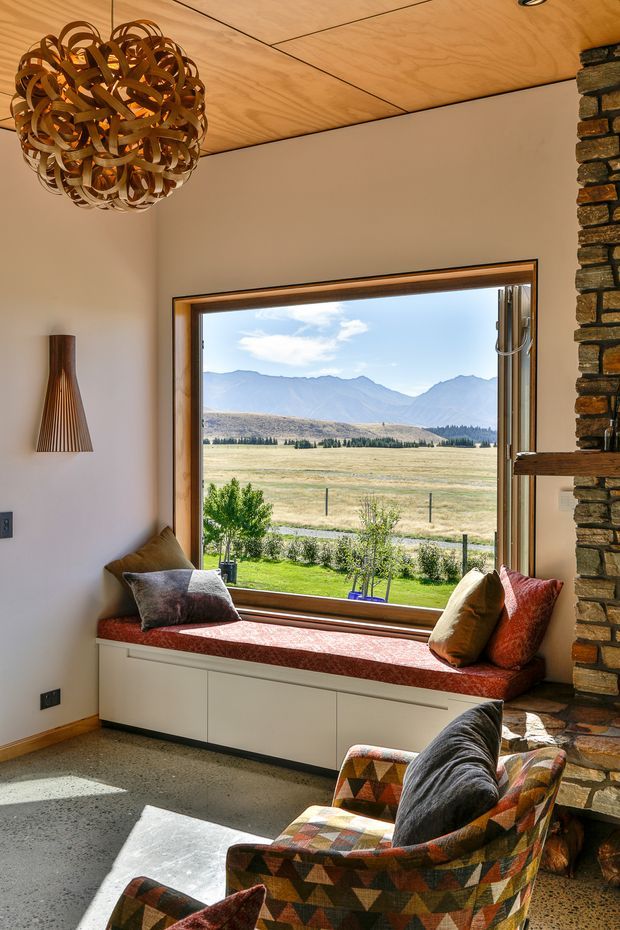Warm, light-filled living room with a large picture window framing a mountain and rural valley landscape. The room features a built-in window seat with red upholstery and cushions, two patterned lounge chairs, a stone fireplace wall, a sculptural woven pendant light, a timber panel ceiling, and polished concrete flooring.
