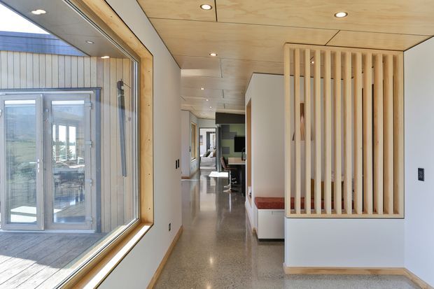 A bright contemporary interior hallway with polished concrete flooring, a light timber-lined ceiling, and a large timber-framed window overlooking an exterior deck. A vertical timber slat screen partially conceals built-in seating or storage, while a kitchen or dining area is visible farther down the corridor.