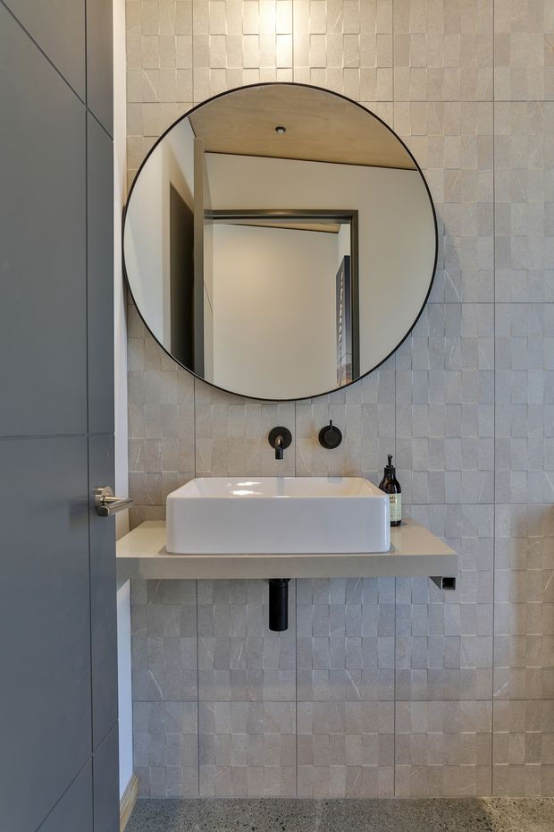 Compact contemporary bathroom vanity area featuring a wall-mounted white vessel basin on a pale stone or engineered-stone shelf, matte black wall-mounted tapware and exposed drain, a large round black-framed mirror, and small-format textured light grey-beige wall tiles. A dark grey door and speckled grey floor create contrast.