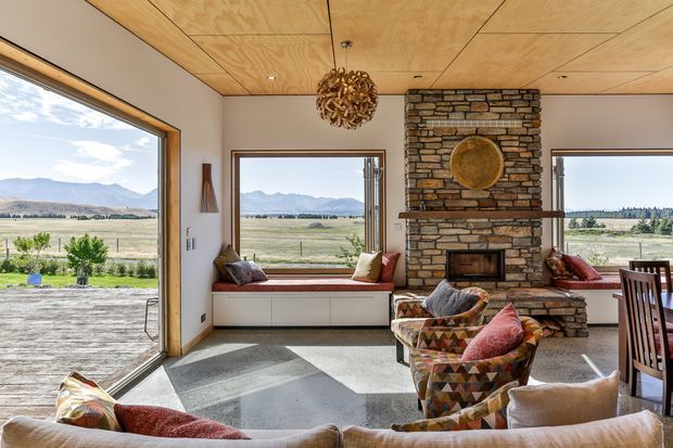A bright, expansive living room with large picture windows overlooking an open rural mountain landscape. The room features a timber-panelled ceiling, polished concrete floor, built-in window seating and cabinetry, a substantial natural-stone fireplace wall, colourful patterned armchairs, and warm contemporary furnishings.