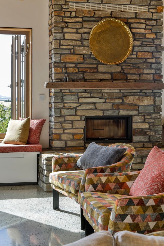 A warm living room centered on a substantial rough-stacked stone fireplace with a timber mantel and inset wood-burning firebox. A large decorative brass-gold round plate hangs above the mantel. Two colourful upholstered armchairs with geometric triangular patterns sit in front of the fireplace, complemented by a window seat with cushions. Daylight enters through the tall window, highlighting the polished speckled concrete floor and natural stonework.