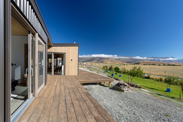 A modern single-storey home viewed from a broad timber deck, with large sliding glass doors, vertical timber-clad walls, and a simple flat-roofed form. The deck overlooks an expansive rural valley with planted grounds, gravel landscaping, and distant rugged mountains beneath a clear blue sky.