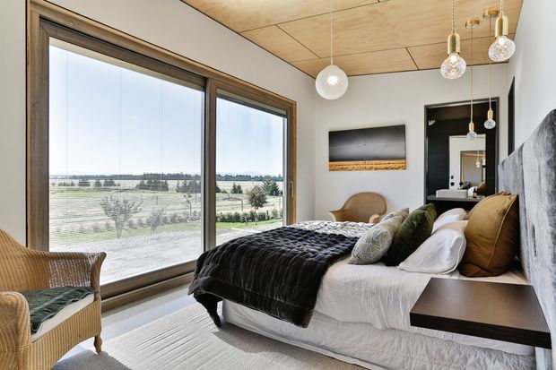 A bright, contemporary bedroom with expansive floor-to-ceiling timber-framed glazing overlooking a rural landscape. The room features a low platform bed with white bedding, a dark textured throw, layered neutral and olive cushions, integrated dark bedside joinery, a textured grey feature wall, a pale timber-lined ceiling, and sculptural pendant lighting. A woven lounge chair sits near the window.