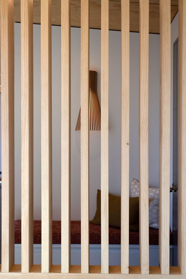 Interior view through a vertical light-timber slatted divider into a compact bedroom or sleeping nook. The space features a raised platform bed with a deep red bedspread, mustard and patterned cushions, white walls, a timber-lined ceiling, a sculptural timber pendant light, and dark-framed glazing or a door at the right. The slatted screen is the dominant architectural feature, creating filtered privacy while allowing light through.