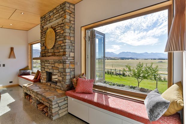 Bright, warm-toned living room with a substantial natural stone fireplace and integrated bench seating beneath expansive timber-framed windows. The room opens to a broad rural landscape with mountains beyond, while red upholstered cushions, a timber ceiling, polished concrete or terrazzo-like flooring, and a sculptural wall light add character.
