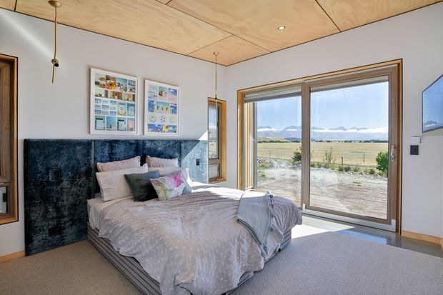 Bright contemporary bedroom with a low bed, dark upholstered feature headboard, framed artwork, pale carpet, and extensive timber-framed glazing opening to a rural landscape. The room has a warm plywood ceiling with recessed lighting and a restrained neutral palette, while a wall-mounted screen is visible at right.