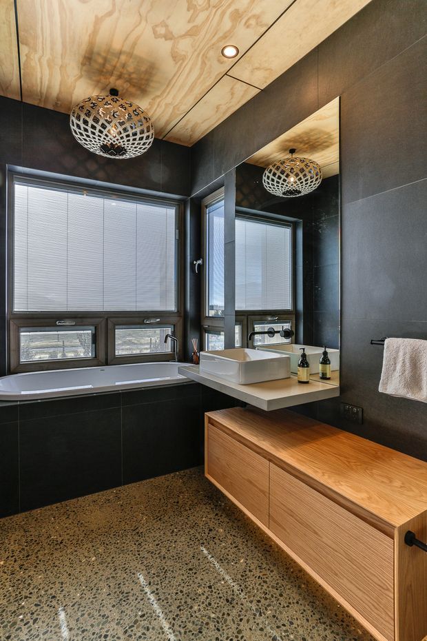 A contemporary bathroom featuring dark large-format wall tiles, a polished exposed-aggregate floor, a timber vanity with drawers, a white vessel basin, and a built-in bathtub beneath wide windows with blinds. The plywood ceiling has recessed lighting, while a decorative woven pendant light is reflected in the large wall mirror.
