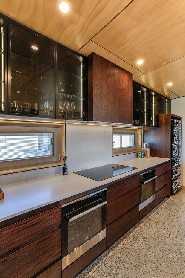 A contemporary galley-style kitchen with dark timber cabinetry, glossy black glass-front overhead cupboards, pale engineered-stone benchtops, integrated black appliances, and a warm timber-lined ceiling. Narrow horizontal windows bring in daylight, while polished concrete or terrazzo-style flooring adds a durable modern finish.