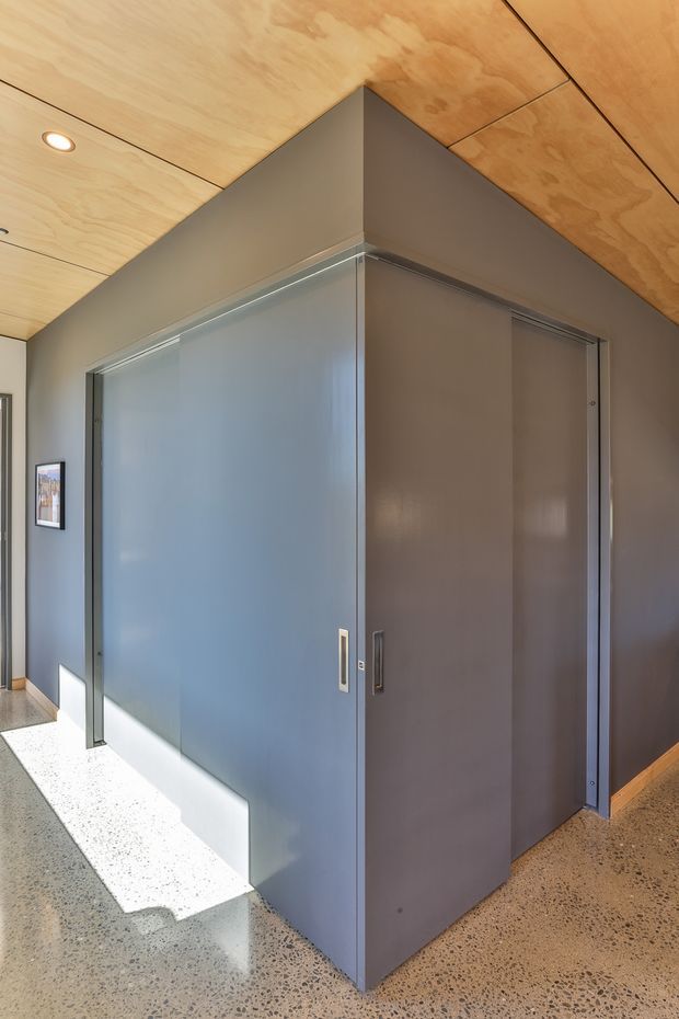 Interior view of a minimalist hallway or entry area featuring a dark grey built-in wall volume with flush sliding doors, a warm plywood-lined ceiling with recessed downlights, polished exposed aggregate concrete flooring, and a small framed artwork.
