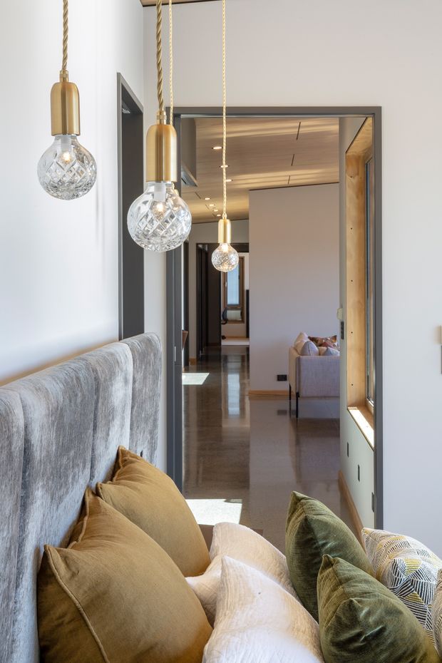 A refined interior view looking from a living area toward a hallway, with a large upholstered sofa and layered neutral cushions in the foreground. Three decorative pendant lights with brass-toned cylindrical fittings and faceted glass globes hang over the seating area. The hallway features polished flooring, dark-framed openings, timber ceiling lining, and a built-in display or storage niche, with another living space visible beyond.