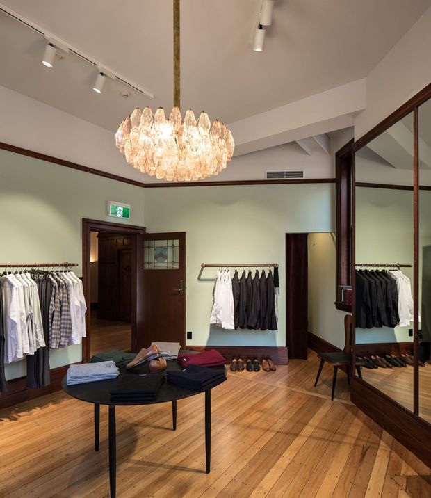 A refined clothing boutique or dressing showroom with timber flooring, pale green walls, dark timber trim and doors, clothing racks, footwear displays, a central round display table, and large mirrored cabinetry. A prominent crystal pendant chandelier hangs beneath a white ceiling with track lighting. The room has a warm, elegant, heritage-inspired retail interior.