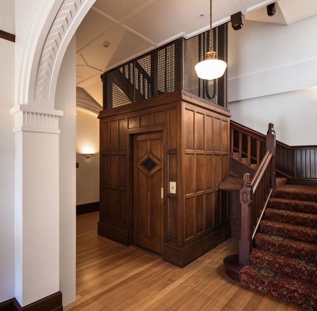 Interior view of a heritage-style stair hall featuring a richly panelled dark timber elevator enclosure, patterned metal screening above it, a carpeted staircase with ornate timber handrails and balustrades, polished timber flooring, and a large white decorative archway. Warm pendant and wall lighting enhance the traditional character.