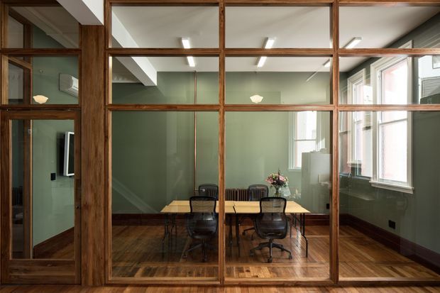 Interior view through a full-height, timber-framed glazed partition into a compact dining and meeting area. The room has muted sage-green walls, dark timber flooring and skirting, a rectangular timber table with office-style chairs, exposed timber glazing frames, and large windows along the right wall. Reflections reveal additional interior space and wall-mounted equipment.
