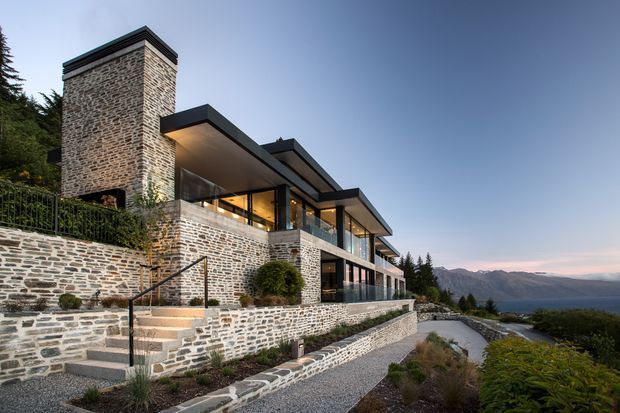 A contemporary multi-level hillside residence with extensive natural stone masonry, dramatic cantilevered flat roof planes, floor-to-ceiling glazing, and stepped terraces. The home overlooks a mountainous landscape and includes landscaped retaining walls, stairs, and a gravel walkway.