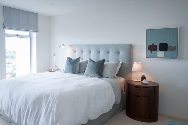 Bright, softly styled bedroom with a large upholstered button-tufted headboard, white bedding, layered grey-blue cushions, and a dark rounded timber bedside cabinet. A floor lamp, table lamp, abstract artwork, and roller blind complete the calm contemporary setting. A window provides natural light and an exterior view.