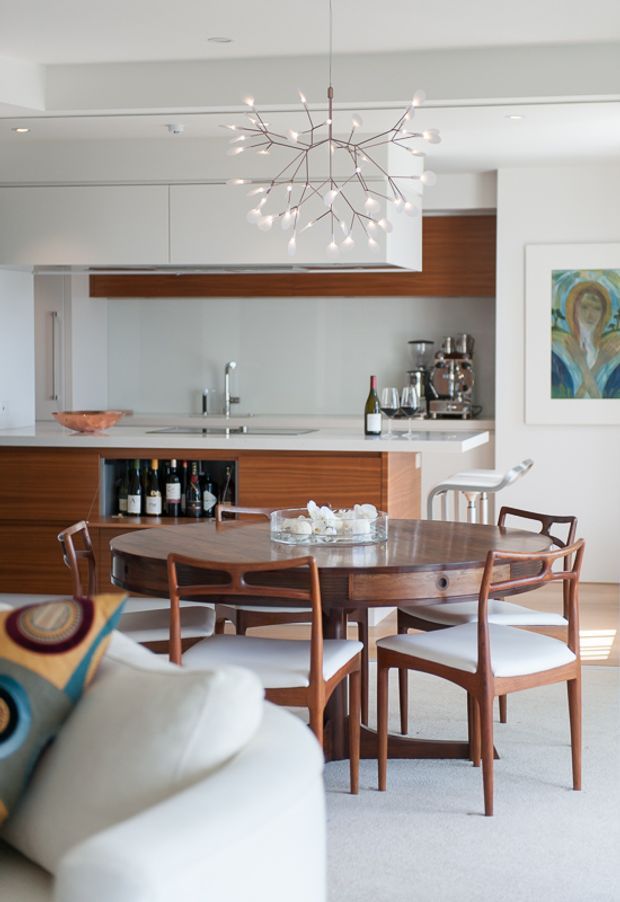 Bright open-plan interior featuring a mid-century-inspired dining area and kitchen. A large oval dark timber dining table with matching timber chairs occupies the foreground, while the background has white cabinetry, a white benchtop, timber cabinetry and splashback detailing, integrated appliances, and a sculptural branching pendant light. A white sofa and colourful artwork are also visible.