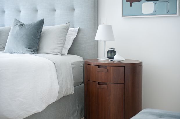 A softly lit bedroom vignette featuring a partially visible upholstered bed with white bedding and pale blue-grey pillows, beside a rounded dark timber bedside cabinet with integrated drawers. A white table lamp, small decorative objects, and abstract artwork complete the calm, refined setting.