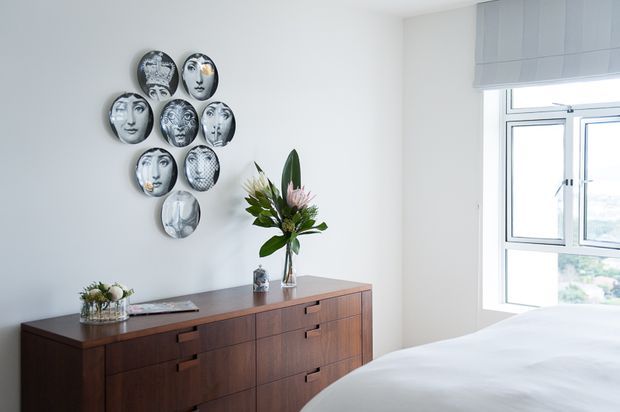 Bright, calm bedroom corner featuring a low mid-century style dark timber credenza, white bedding, a large window with a light roller blind, and a decorative arrangement of monochrome face plates above the cabinet. A vase of tropical foliage and flowers adds a natural accent.