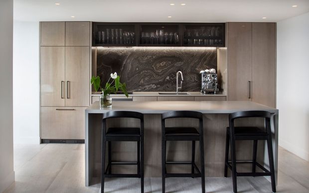 Contemporary kitchen featuring a large central island with a dark stone-look benchtop, three black bar stools, integrated timber-toned cabinetry, open glassware shelving, under-cabinet lighting, a sculptural dark marble-look splashback, and a stainless-steel sink and tap. The space has a clean, refined, neutral palette with recessed ceiling lighting.