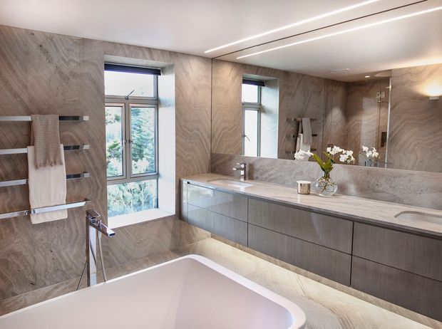 A spacious contemporary bathroom featuring a freestanding white bathtub, long double vanity with integrated sinks, large wall-to-wall mirror, glass-enclosed shower, heated towel rails, and full-height veined stone-look wall and floor finishes. Linear recessed ceiling lighting and under-vanity illumination create a refined, softly lit interior.