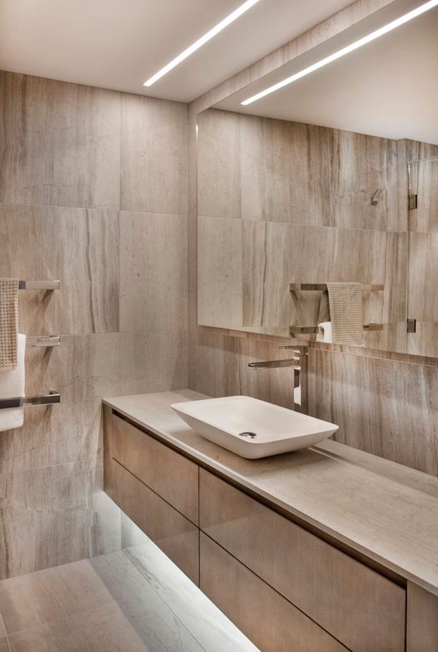 Contemporary bathroom with floor-to-ceiling beige stone-look tile walls and flooring, a long floating timber-toned vanity, white vessel basin, minimalist chrome tapware, large wall mirror, integrated shelving, and linear recessed ceiling lighting.