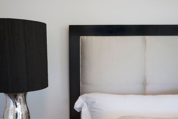 Close-up of a refined bedroom vignette featuring a white upholstered bedhead framed in dark timber, crisp white bedding, and a black textured drum table lamp with a reflective metallic base against a light wall.