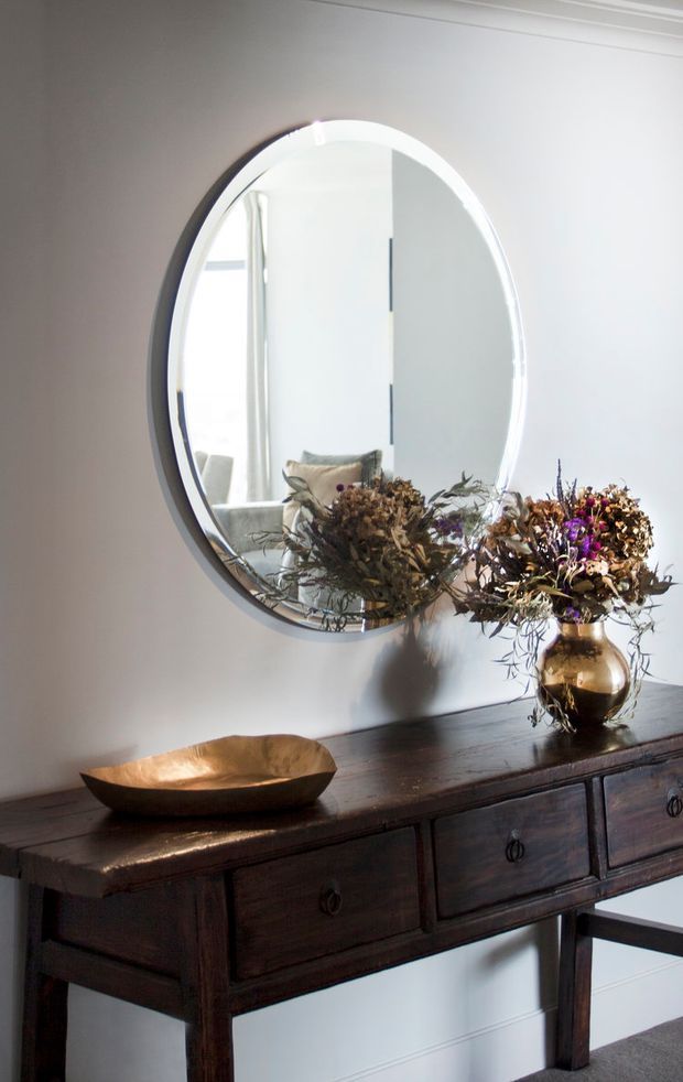 A bright, refined interior vignette featuring a large round wall mirror above a dark timber console table. The console has multiple drawers and a substantial wood top, styled with a shallow metallic bowl and a brass-toned vase holding dried flowers. White walls with simple cornice detailing and a glimpse of upholstered seating are reflected in the mirror.
