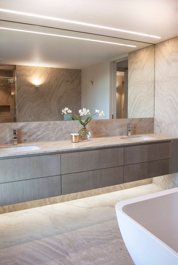 Contemporary luxury bathroom featuring a wide double vanity with integrated basins, dark wood-toned floating cabinetry, pale veined stone benchtop and wall surfaces, a large mirror with linear integrated lighting, and a freestanding white bathtub in the foreground. A glass shower enclosure is visible at left, with understated floral styling on the vanity.